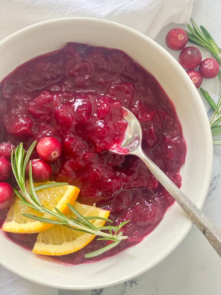Close up photo: cranberry sauce in a bowl with orange, cranberries, and rosemary as garnish.