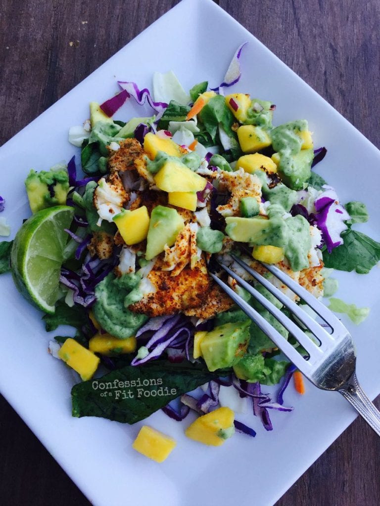 Fish taco salad with red cabbage, diced mango, diced avocado, taco seasoned fish, and greens on a square white plate and a fork poised for eating