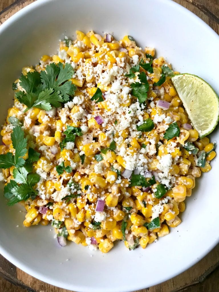 A white bowl filled with Mexican street corn salad topped with cotija cheese and cilantro with a lime wedge on the side