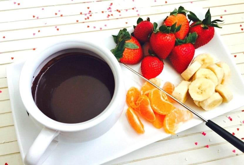 A small white pot of dairy free chocolate fondue on a white rectangular serving dish. On the dish are whole strawberries, sliced bananas, and mandarin orange segments. There are small pieces of heart shaped confetti on the white wooden background.