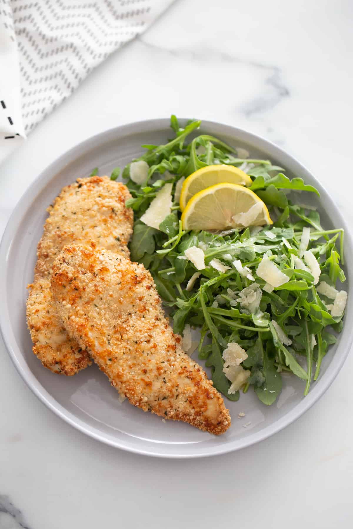 Overhead shot of golden brown two chicken cutlets plated with a side salad of arugula, lemon, and shredded parmesan