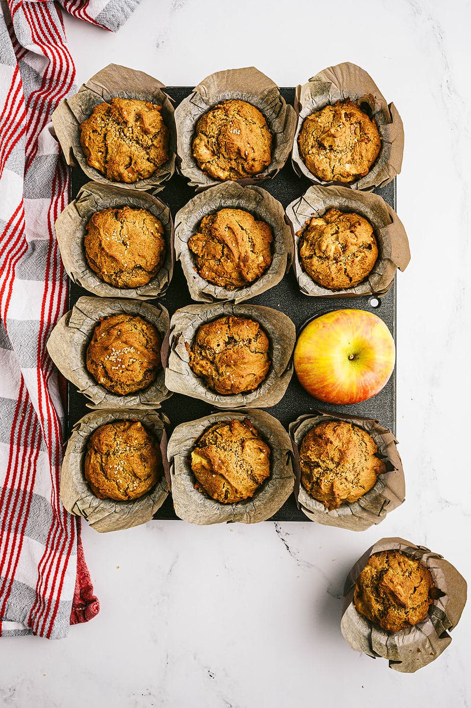 Overhead photo: muffin tin full of cooked muffins with a whole apple taking one's place.