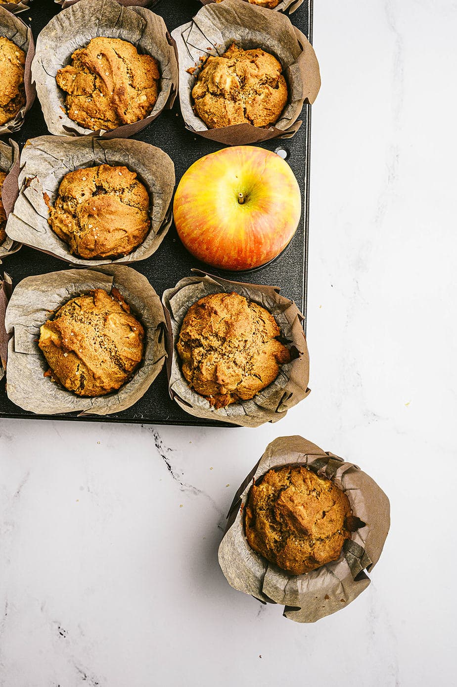 Overhead image: parchment lined muffins in a muffin tin. One muffin is out of its place, off to the side and a whole apple takes its place in the tin.