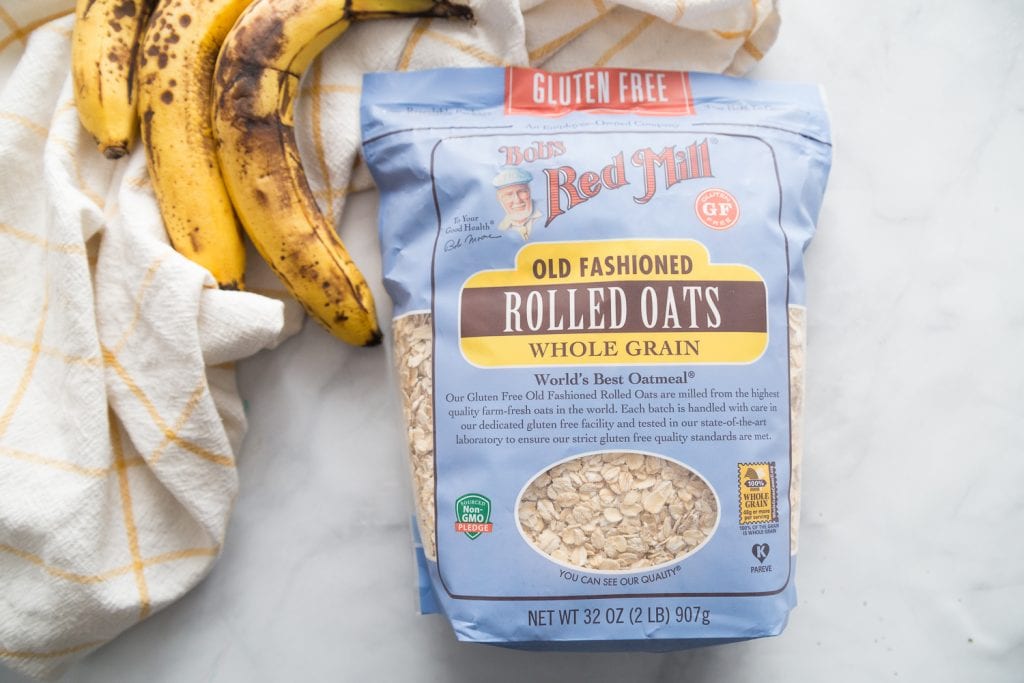 A bag of Bob's Red Mills Gluten Free Oats and three very ripe bananas sit on a white marble backround