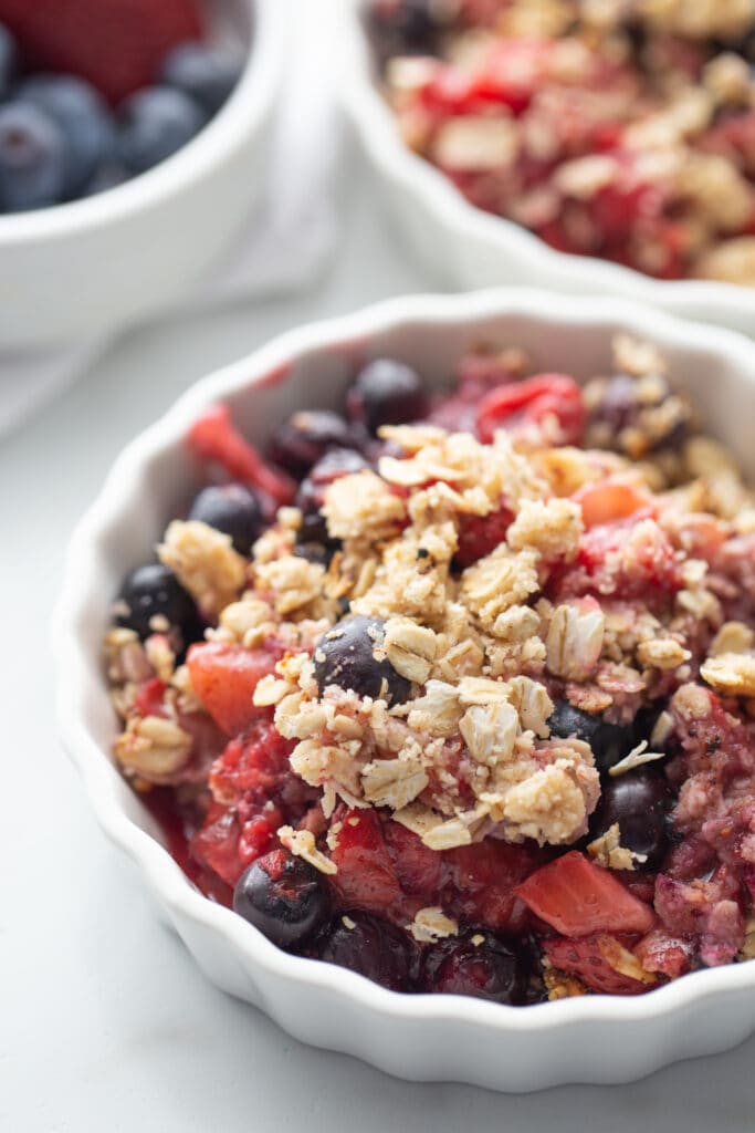 Mixed berry crisp with gluten free oat topping in shallow ramekin. In the background, out of focus, are two more white ramekins of blueberry crisp with strawberries and raspberries.. 