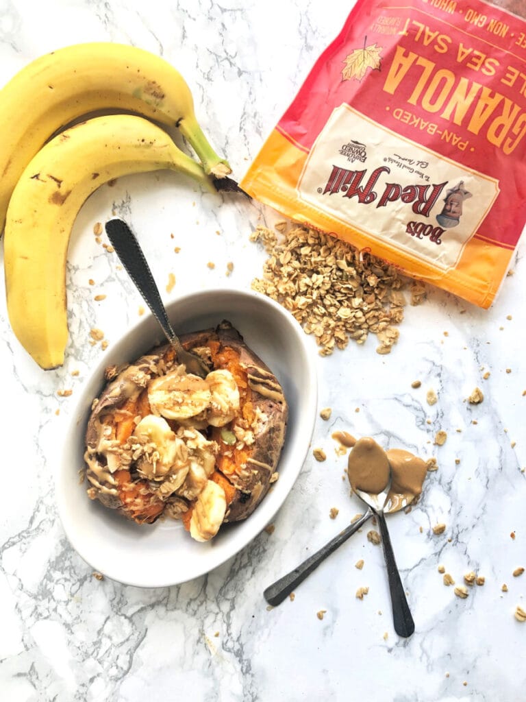 A bowl of sweet potato stuffed with banana and peanut butter on a marble counter. Bananas and a bag of Bob's Red Mill Maple Sea Salt granola are sitting on the counter nearby 