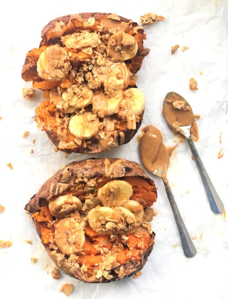 A photo of stuffed sweet potatoes on a marble background stuffed with peanut butter, bananas, and granola. Two spoons of peanut butter sit nearby