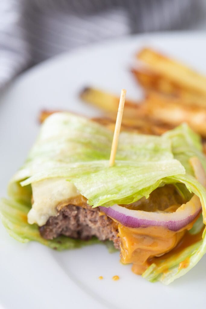 A toothpick is holding together a lettuce wrapped burger with delicious toppings and burger sauce, with homemade french fries in the background.