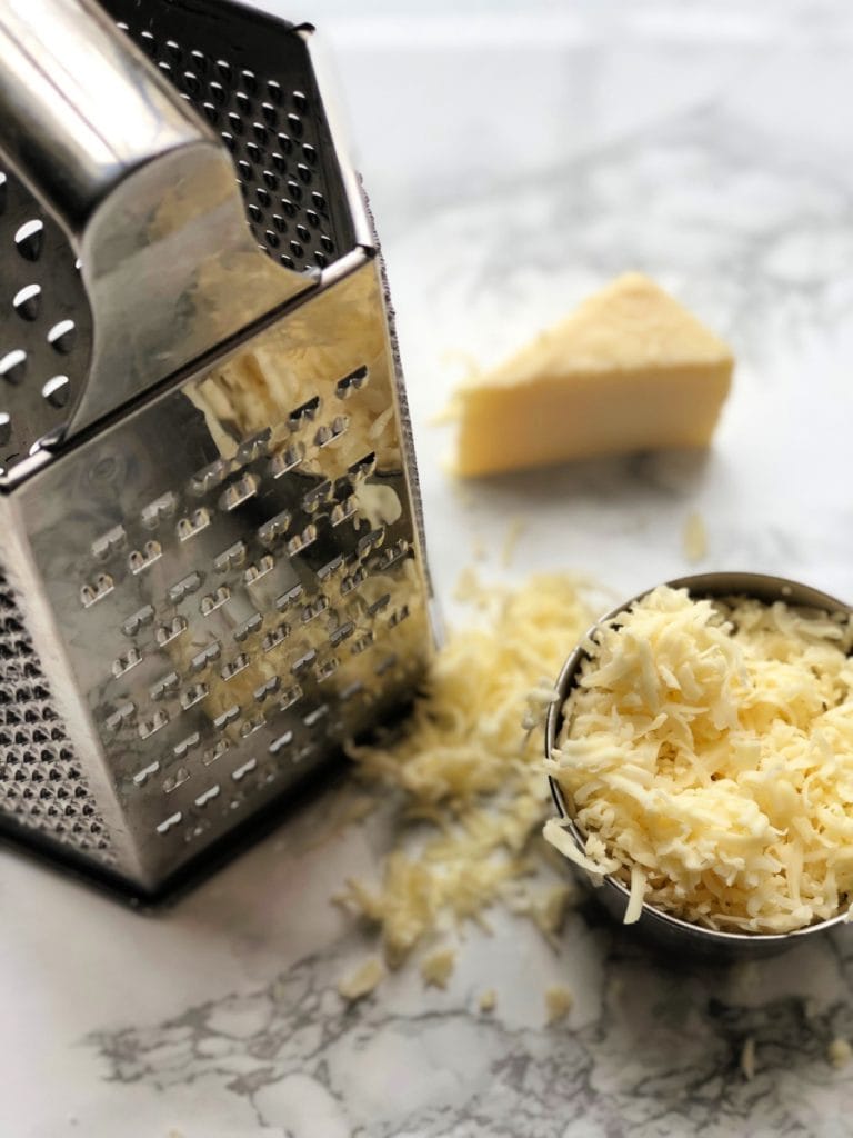 A box grater is sitting next to a cup of shredded cheddar on a marble background. A triangle piece of cheese sits behind. 