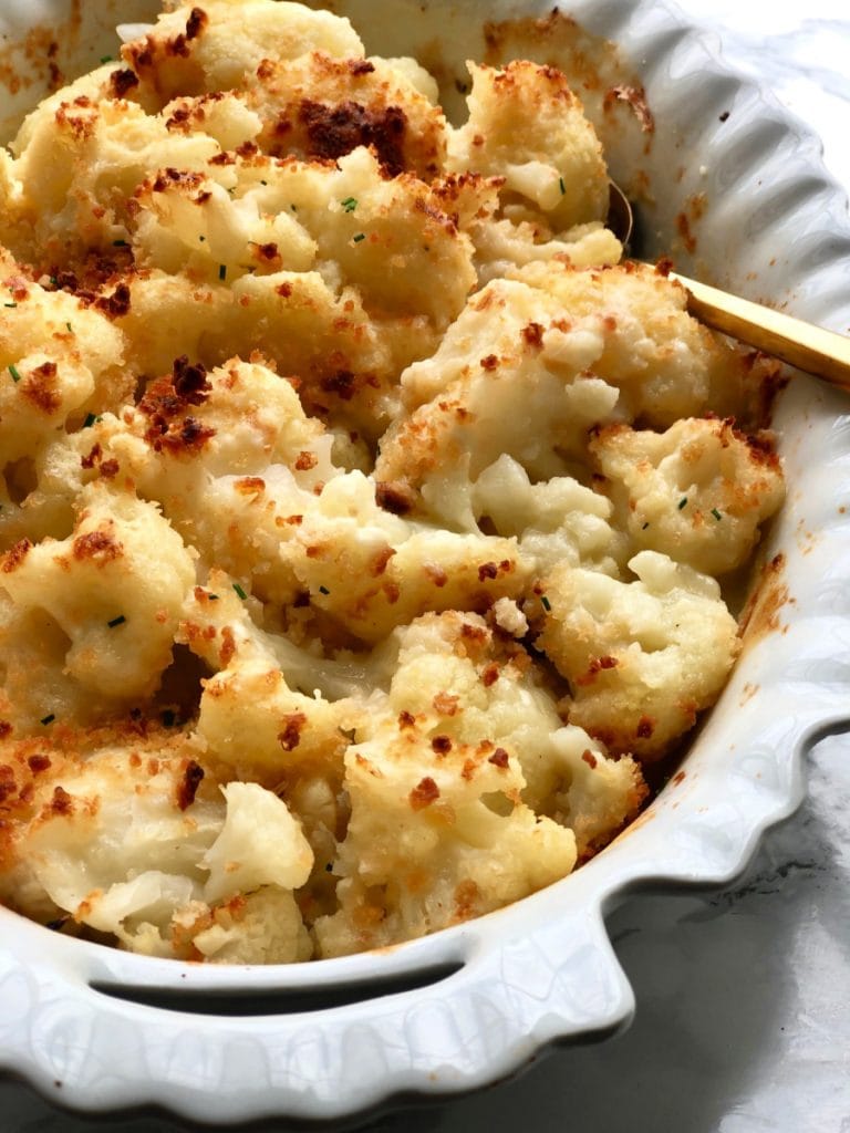 A freshly baked cauliflower au gratin bake sits on a white marble background with a gold serving spoon 