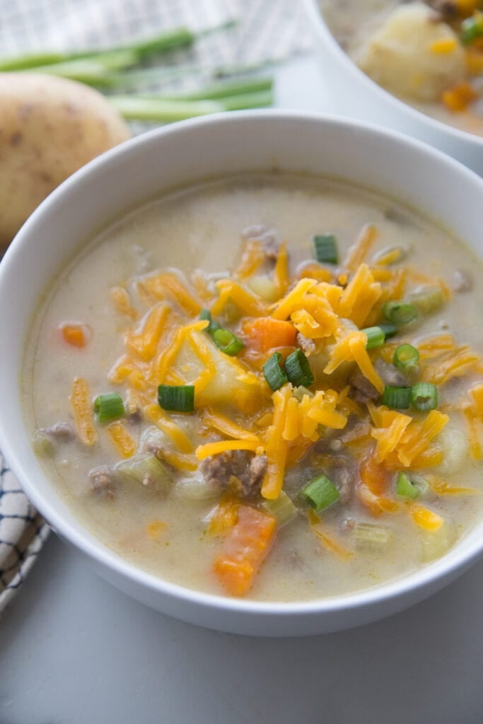 A bowl full of creamy Cheeseburger Soup topped with cheddar cheese and green onions sitting on a white table with a second bowl in the background