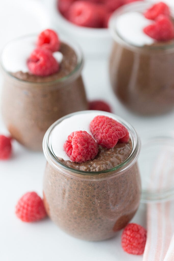 Photo of Chocolate chia pudding in a glass cup topped with whole raspberries and coconut cream. There are more raspberries and glass containers full of chocolate chia pudding in the background, out of focus.