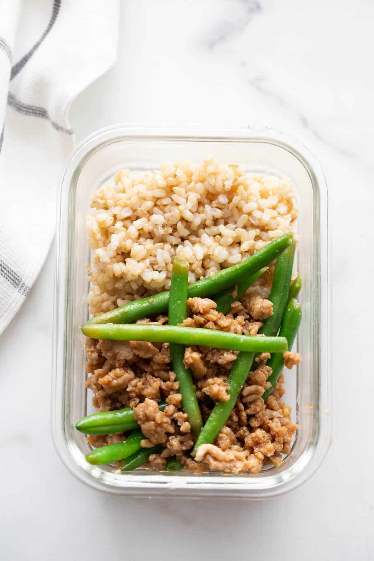 A glass meal prep container with brown rice and ground chicken stir fry and green beans.
