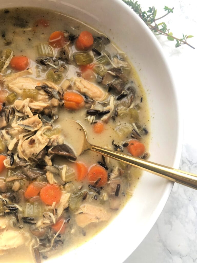 Overhead photo of a white bowl full of Instant Pot Chicken and Wild Rice Soup with a gold spoon on the side and a white marble background. In the soup there are diced carrots, celery and mushrooms, shredded chicken, wild rice, and a creamy broth.