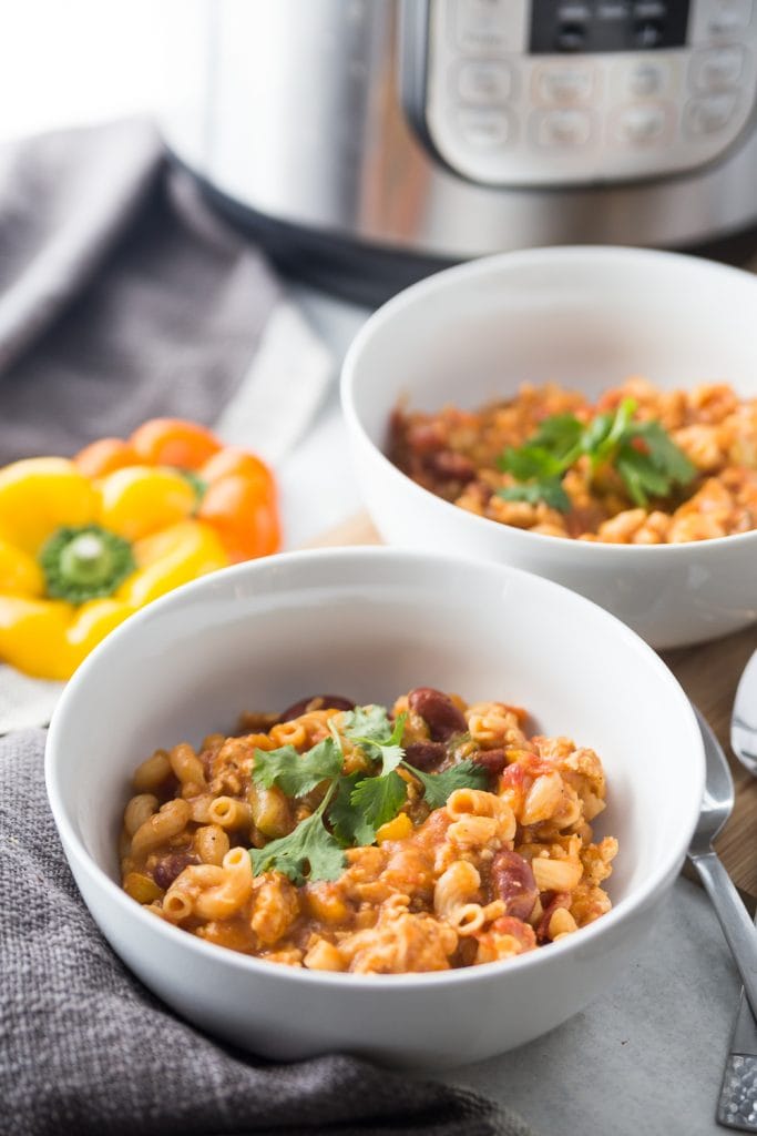 Two bowls of healthy instant pot chili mac sit together in front of an Instant Pot. Two bell peppers sit nearby. 
