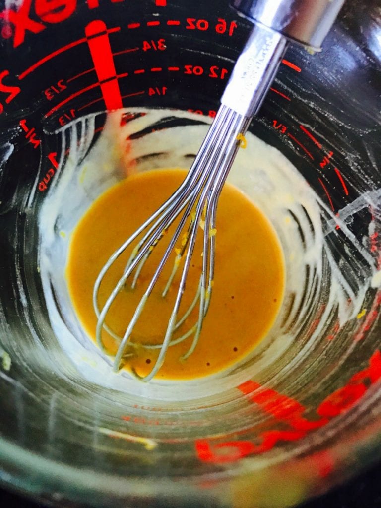 Overhead photo of a glass measuring bowl with a metal whisk mixing citrus maple dijon dressing to top turkey burgers.