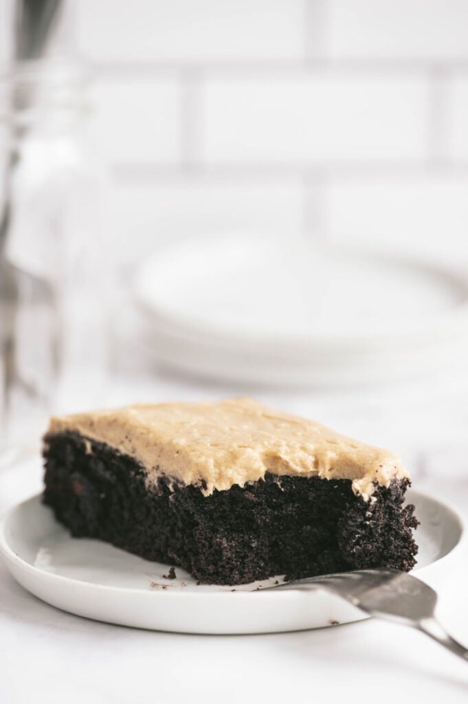 Close up side view of a rectangular piece of Chocolate cake topped with peanut butter frosting on a round white plate with a fork poised upside down on the rim.