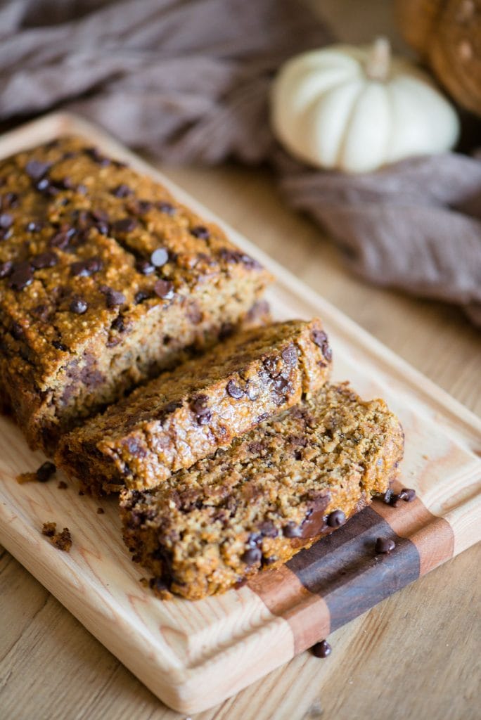 Sliced Pumpkin Oatmeal bread with chocolate chips laying on a wooden cutting board. The light colored wood table is adorned with white and orange pumpkins and a light brown piece of fabric. 
