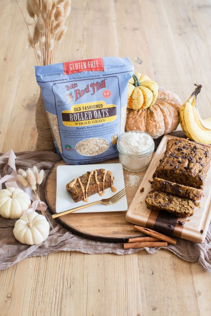 A bag of Bob's Red Mill Old Fashioned Gluten Free Rolled Oats sits in the middle of a festive Autumn table with white, yellow, and orange pumpkins and decorative wheat. A piece of banana bread and peanut butter is on a white plate and a wooden cutting board sits nearby by the full loaf of bread. 