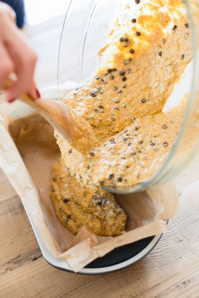 A woman's hand is shown using a wooden spoon to pour oatmeal banana pumpkin bread into a loaf pan lined with parchment paper. 
