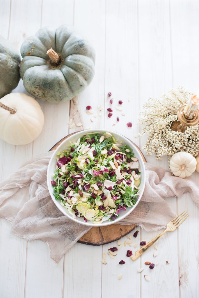 Overhead photo of Maple Cider Holiday Salad with festive fall decor. White, cream, and muted blue decorations surround the white serving bowl full of greens, dried cranberries, and sliced almonds.