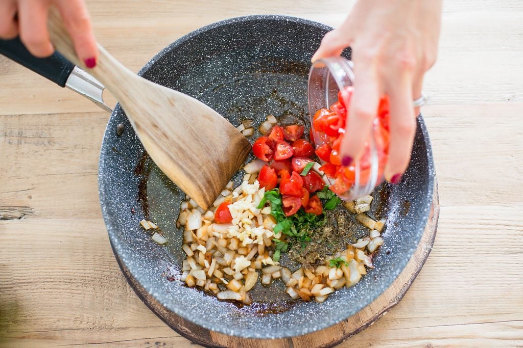 A woman is mixing tomatoes, garlic, onions, and basil in a pan on a wooden table. 