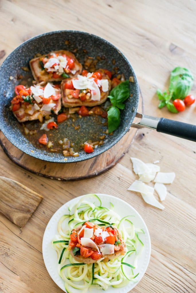 Skillet with three Italian Pork Chops topped with tomatoes, fresh basil, garlic, onions and parmesan cheese. A small white plate is nearby with a pork chop over zucchini noodles. 