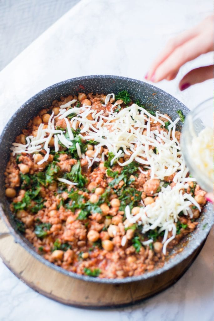 Cheese being placed on top of sausage bean and kale skillet 