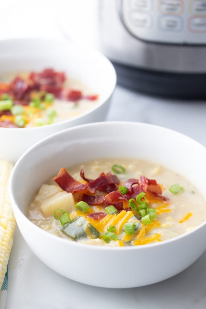 Two bowls of corn chowder topped with bacon, cheese, and green onions sit on a white table in front of an instant pot brand pressure cooker