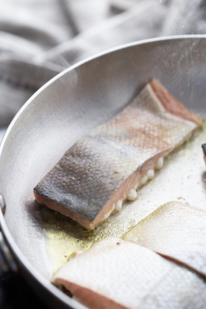 Salmon cooking skin side up in a stainless steel pan.