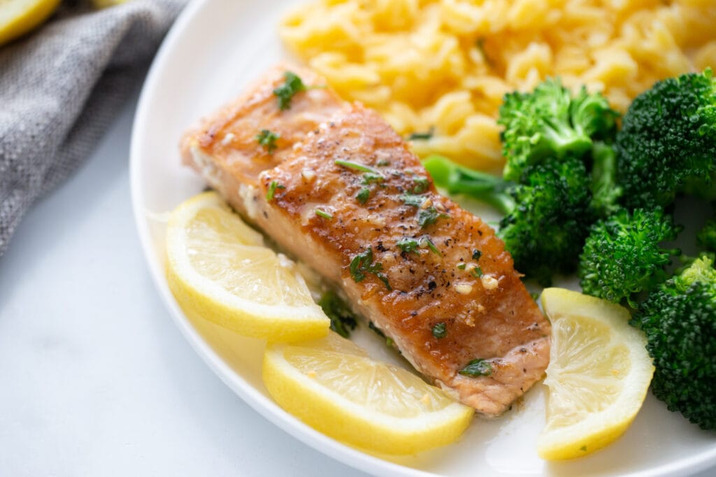 Pan seared salmon on a white plate with sliced lemons and sides of broccoli and gluten free orzo.