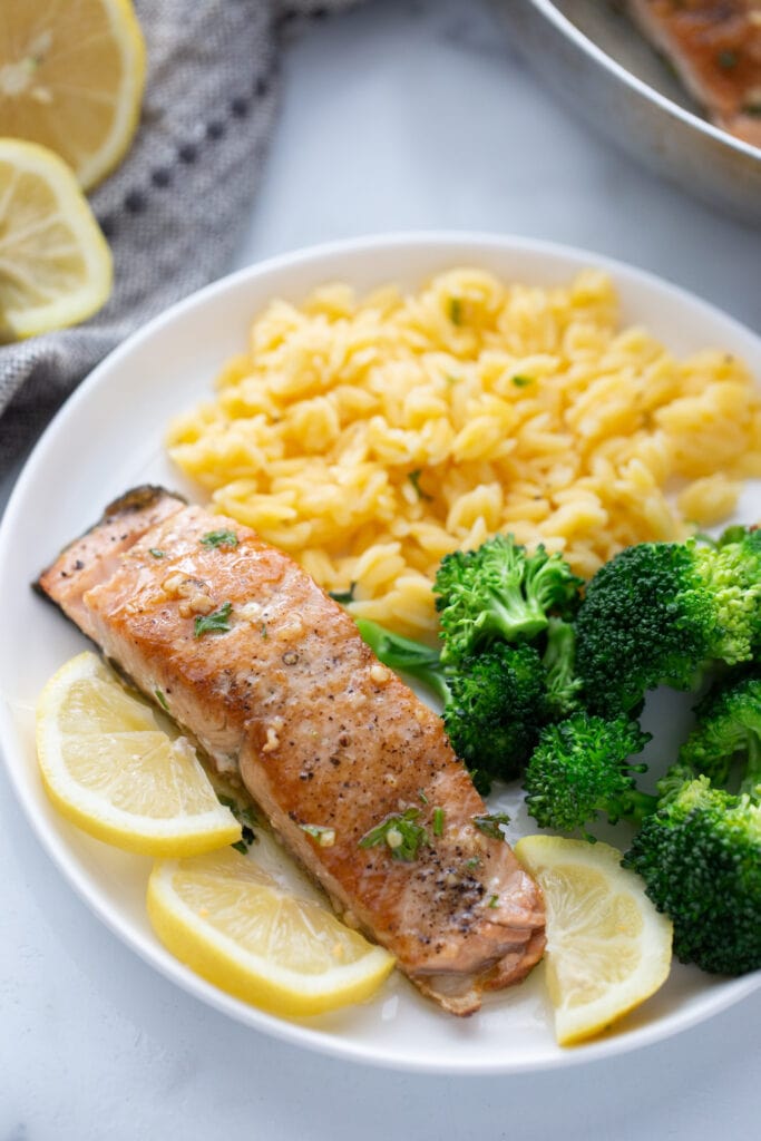 High angle photo of a crispy salmon fillet on a white plate with broccoli and gluten free orzo.