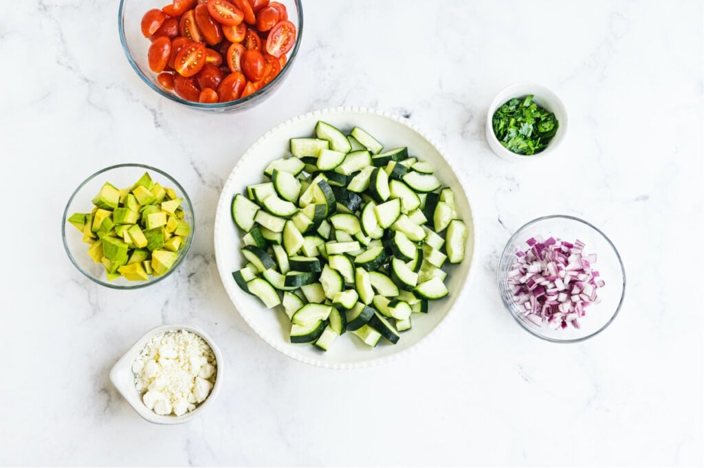 Individual white and clear bowls of ingredients to make cucumber tomato salad on a white background.