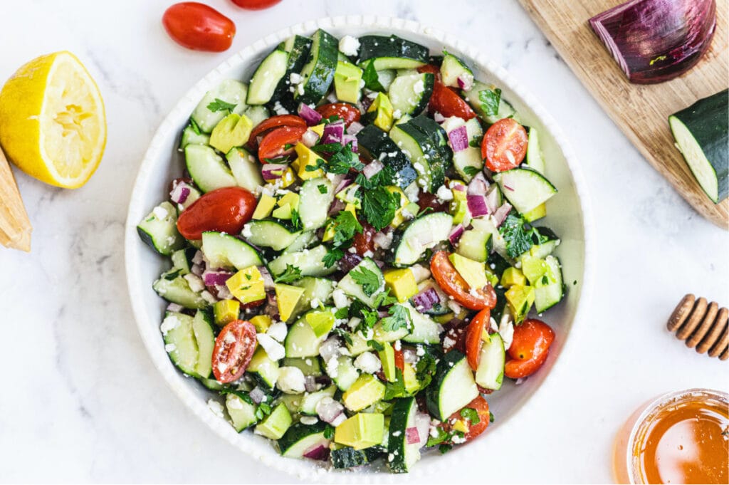 A large white bowl of summer cucumber tomato salad topped with avocado and feta cheese with other ingredients on the side of the bowl.