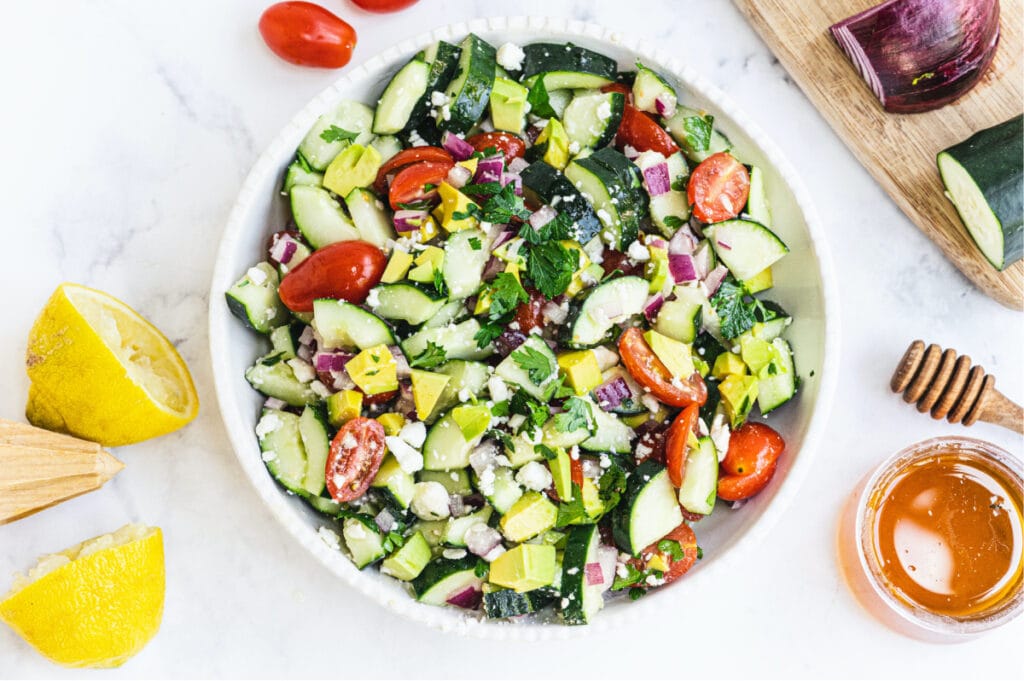Salad made of tomatoes, cucumbers, red onion, feta, and avocado in a large white serving bowl. Lemons, honey, tomatoes, red onion, and cucumber all surround the photo at the edges.