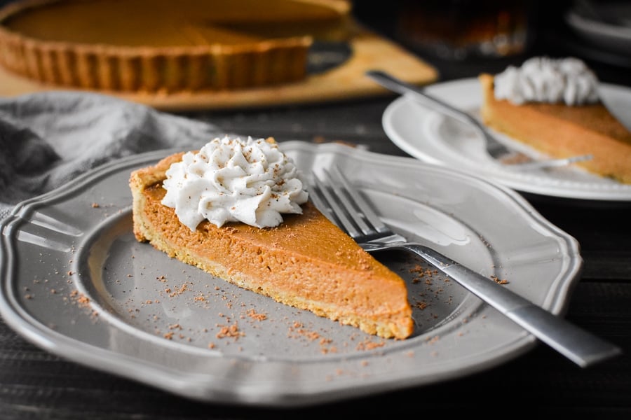 Slice of Pumpkin custard tart with whipped topping on gray plate with whole tart in the background