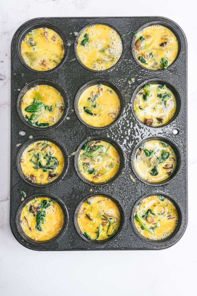 Whisked eggs have been poured on top of the veggies and bacon in the muffin tins.