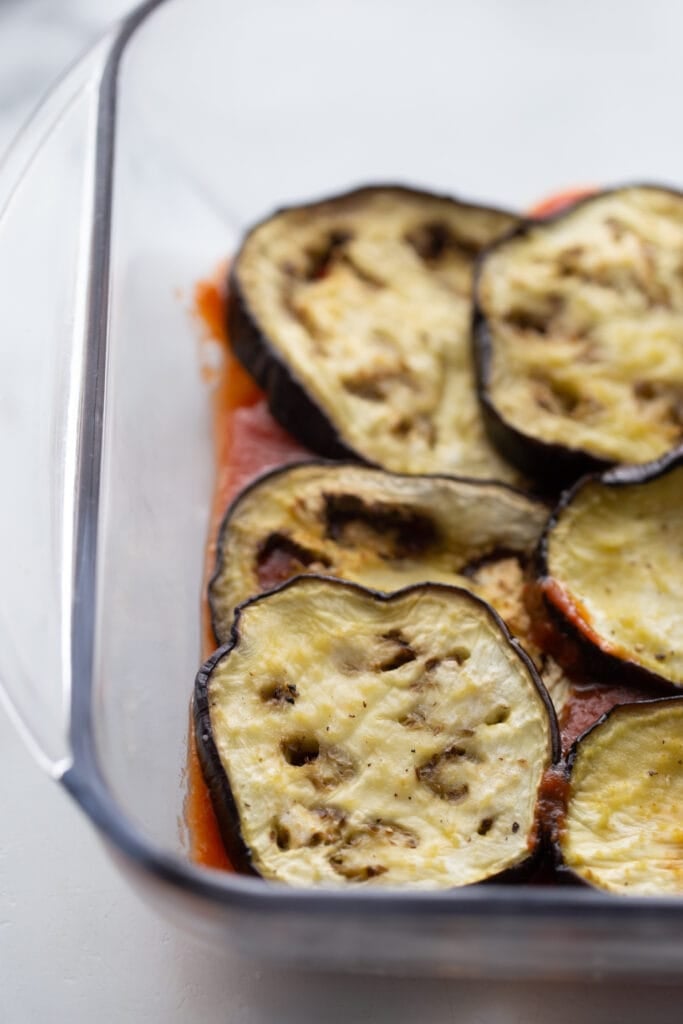 Eggplant slices are placed on top of tomato sauce in a glass baking dish.