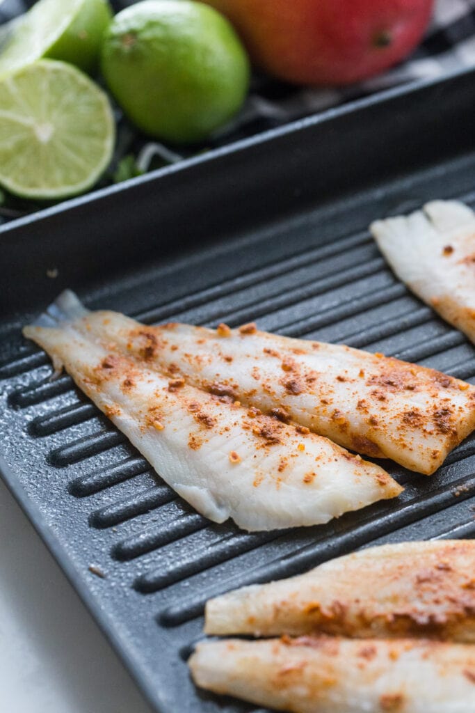 Seasoned White fish filets on a grill pan, ready to be cooked for fish tacos. Limes and mangoes are in the background.