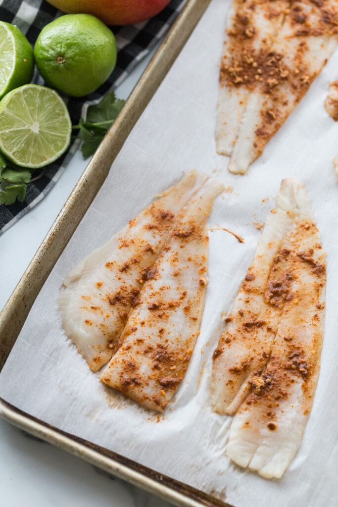 Seasoned white fish filets, on a parchment lined sheet pan, ready for the oven, with fresh limes on the side of the pan.