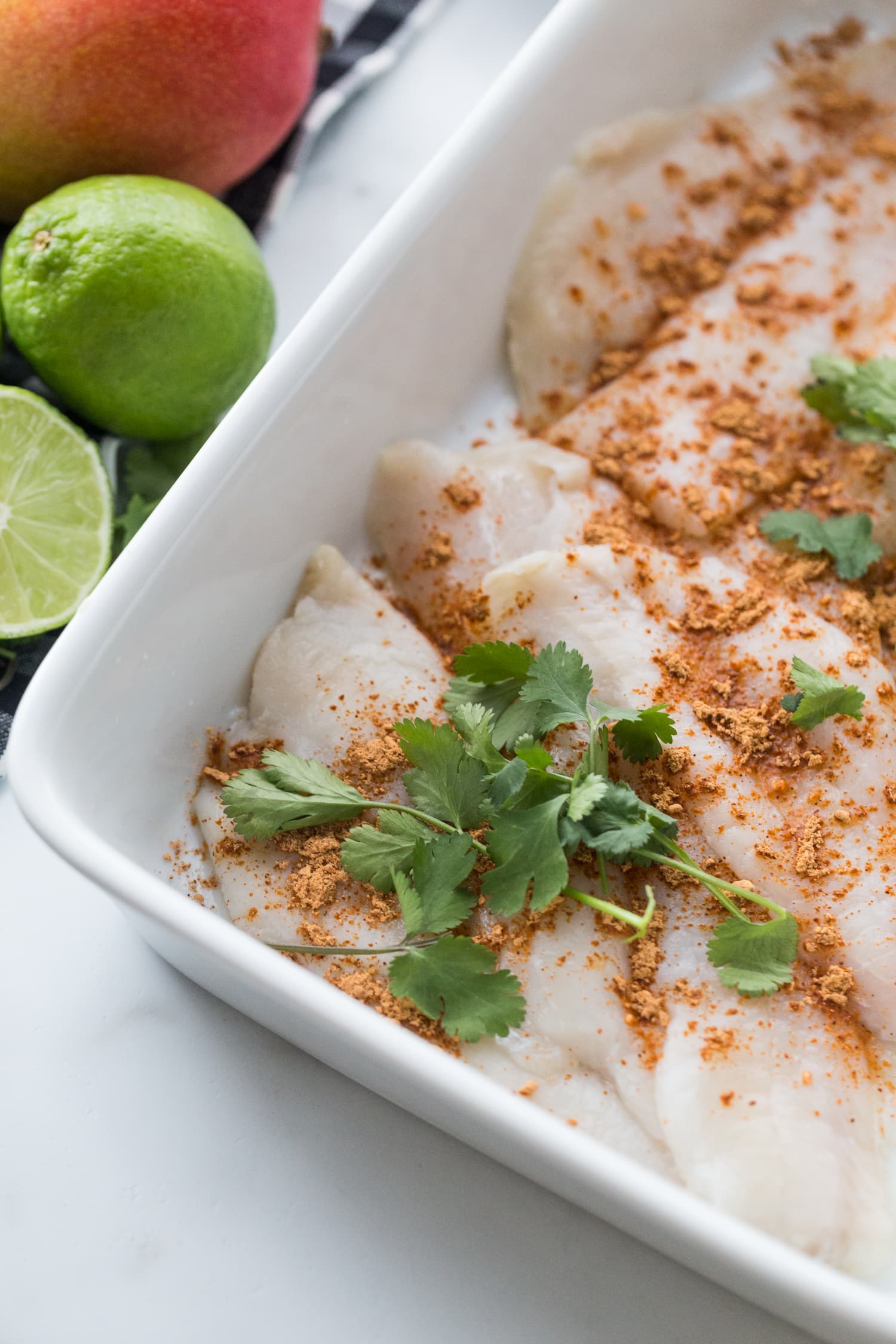 White fish in a white baking dish, topped with spices and cilantro ready to be cooked for fish tacos.