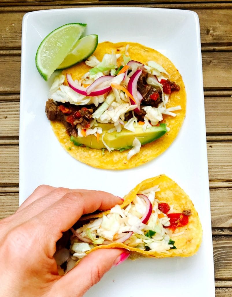 Two Instant Pot Flank Steak Tacos are side by side on a white plate with limes on the side and on a wooden surface. A hand is holding one of the tacos, ready to eat. In the tacos are flank steak, cilantro lime slaw, and topped with cilantro and avocado.