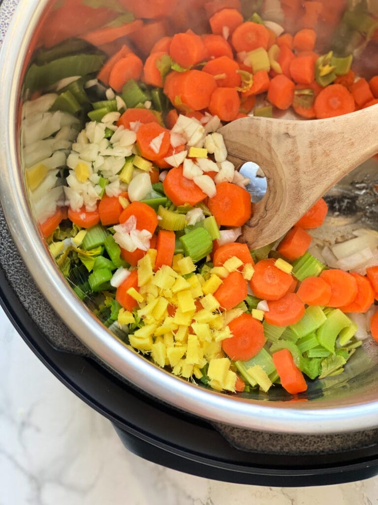 Overhead photo of sliced carrots, celery, onions, garlic, and fresh ginger being sauteed in an Instant Pot, the beginning of Chicken Noodle Soup.