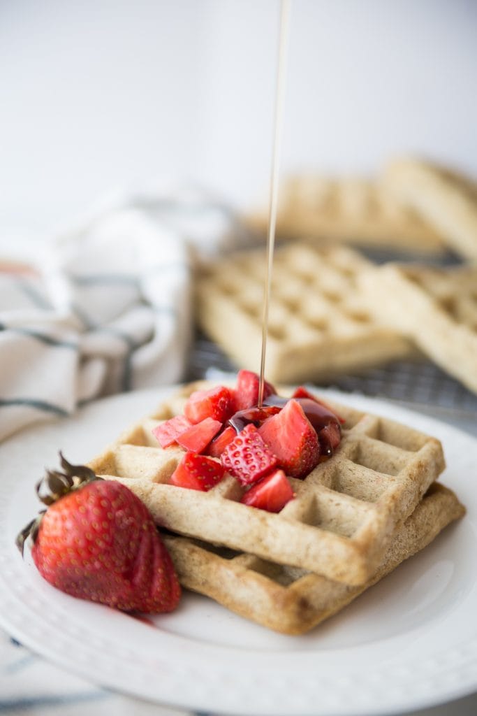 Two square homemade waffles on a white plate topped with diced strawberries and a sliced strawberry for garnish. Maple syrup is being drizzled from above.