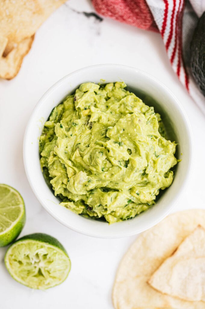 A bowl of guacamole sitting on a white background with limes and chips nearby