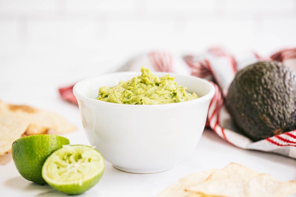A white bowl of homemade guacamole with a chips, some fresh lime and avocado the background