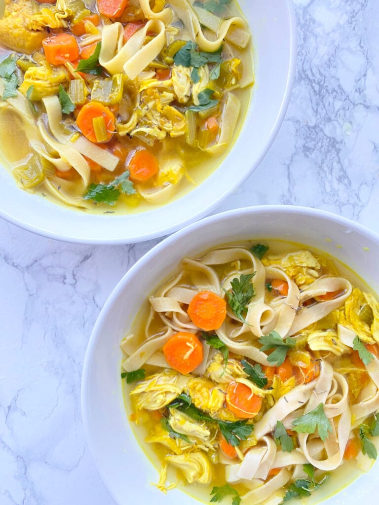 Overhead photo of two hearty bowls of Chicken Noodle Soup filled with sliced carrots, cilantro, egg noodles, and chicken with a beautiful yellow broth colored by Turmeric,