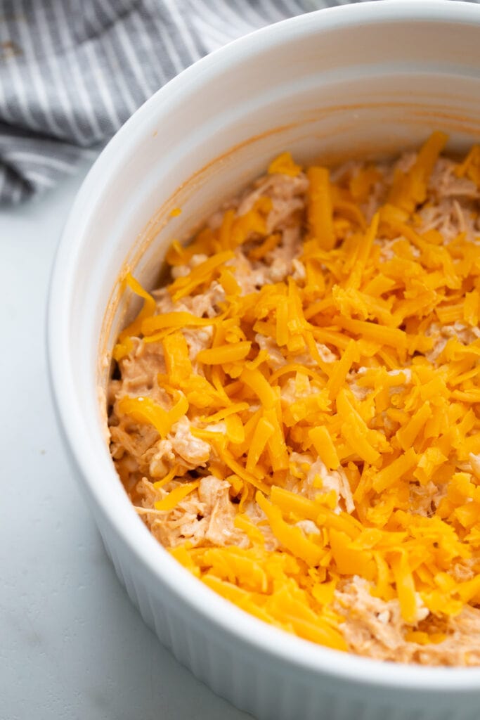 White casserole dish with buffalo chicken dip and topped with shredded cheddar cheese, ready for the oven.