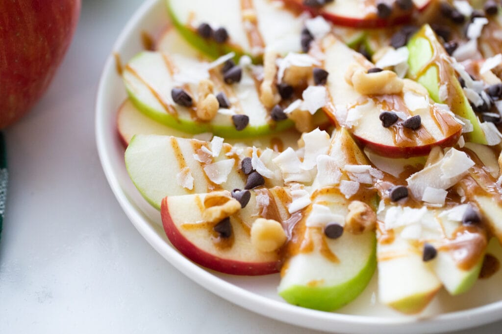 Close up photo of a plate of apple nachos- sliced red and green apples topped with shredded coconut, mini chocolate chips, walnuts, and caramel sauce.