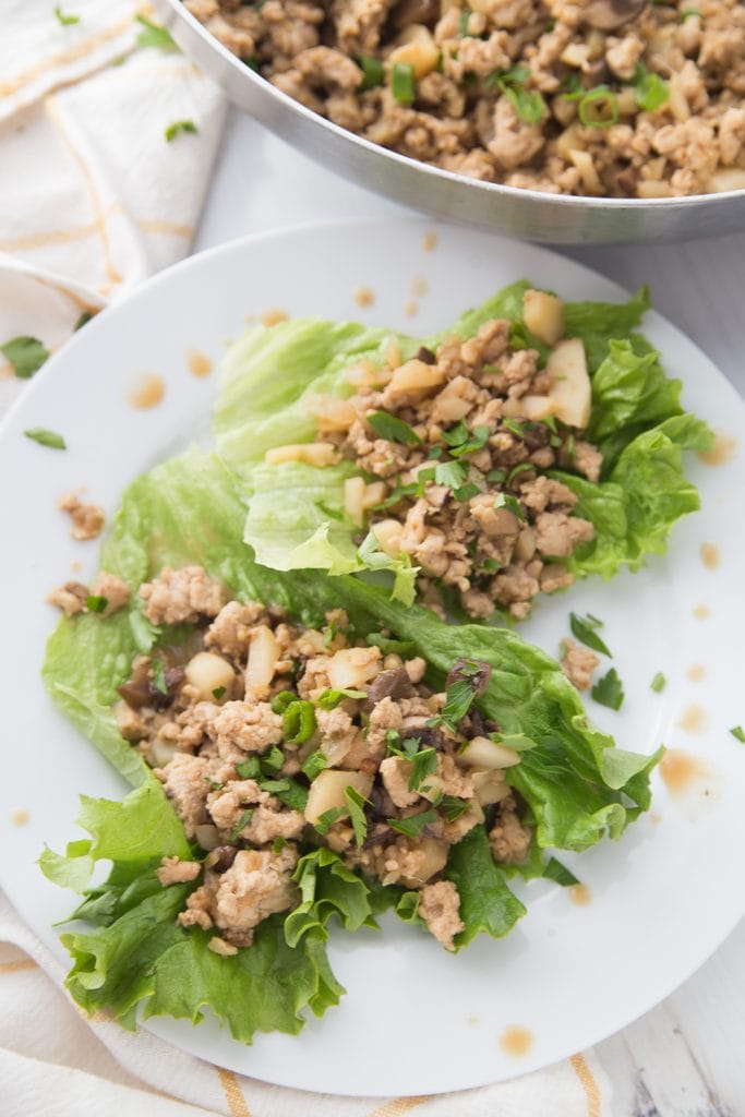 Ground chicken and veggie Lettuce wraps laying on a white plate with a pan of filling in the background
